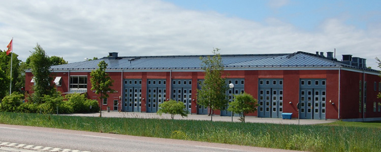 Bärby brandstation - en rödaktig byggnad med gråblå garagedörrar. I förgrunden finns en gräsmatta och några träd.