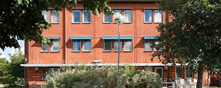 Fålhagsleden 59 0ch 61 - en röd tegelbyggnad med blå fönstermarkiser. En häck och två träd syns i förgrunden.