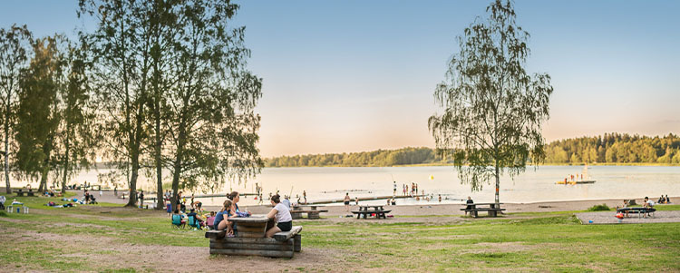 Björklinge friluftsområde - en badplats med sandstrand och bryggor där folk badar och sitter vid bänkar och bord. 