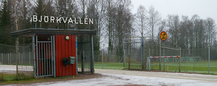 Stängsel och entrén till Björkvallens idrottsplats. I bakgrunden finns en fotbollsplan och träd.