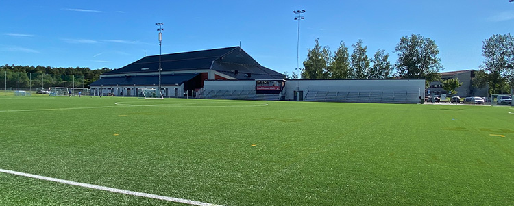 Lötens sportfält - det är en konstgräsplan och i bakgrunden finns träd och en fotbollshall i mörkgrått.
