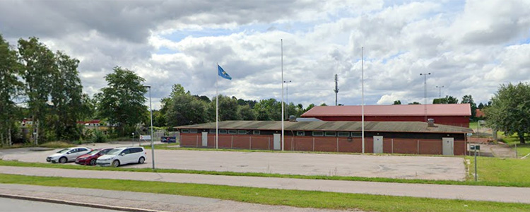 Valsätra idrottsplats, en brun byggnad med staket och tre flaggstänger framför. I förgrunden finns en parkering.